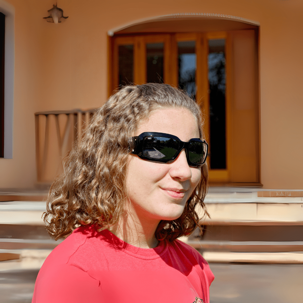 Foto de mulher com óculos de Sol, blusa cor goiaba em frente a uma porta grande de madeira em um casarão.