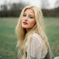 Mulher loira, cabelos soltos, blusa branca em campo aberto gramado.